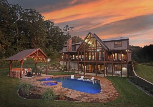 Modern two-story house with a large glass-front, stone chimney, and a stone-paved pool area at dusk.